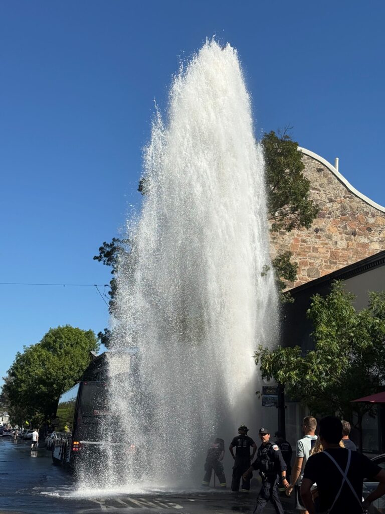 A tall plume of water sprays from a broken fire hydrant over Main Street in St. Helena, with a bus at left and firefighters at the base.