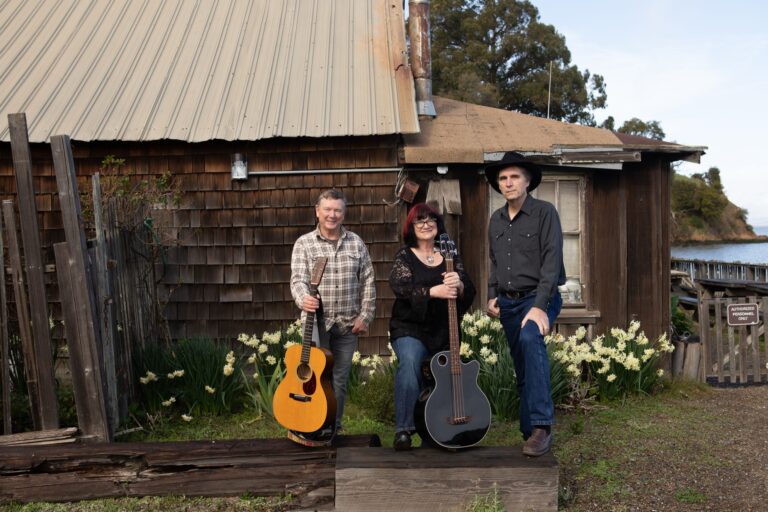 The Larkdales, three musicians with an acoustic guitar and a bass guitar, in front of a weathered shingled cabin with daffodils