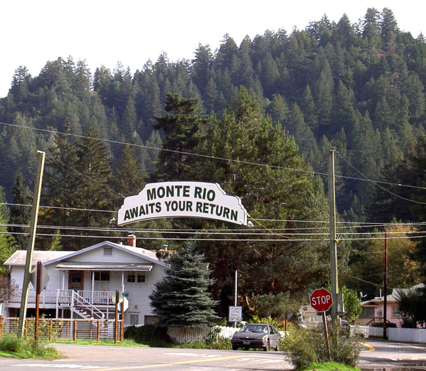 Sign reading Monte Rio Awaits Your Return over a street with a house and wooded hills behind